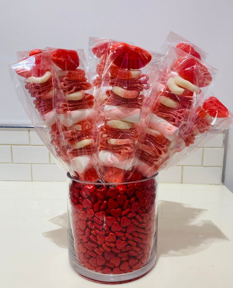 Op de afbeelding: Een glazen vaas gevuld met rode hartvormige snoepjes. Vijf heldere, in plastic verpakte snoepspiesjes zijn in de vaas gerangschikt. Elke spies is bedekt met een rood snoepje in de vorm van een lippenpaar. De spiesjes zijn gevuld met rood en wit snoep.