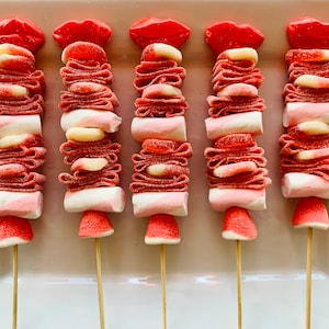 Op de afbeelding: Vijf roze en witte snoepspiesjes met rode snoeplippen erop. Elke spies heeft een marshmallow, roze en witte snoepjes en een roze snoepje in de vorm van een hart.