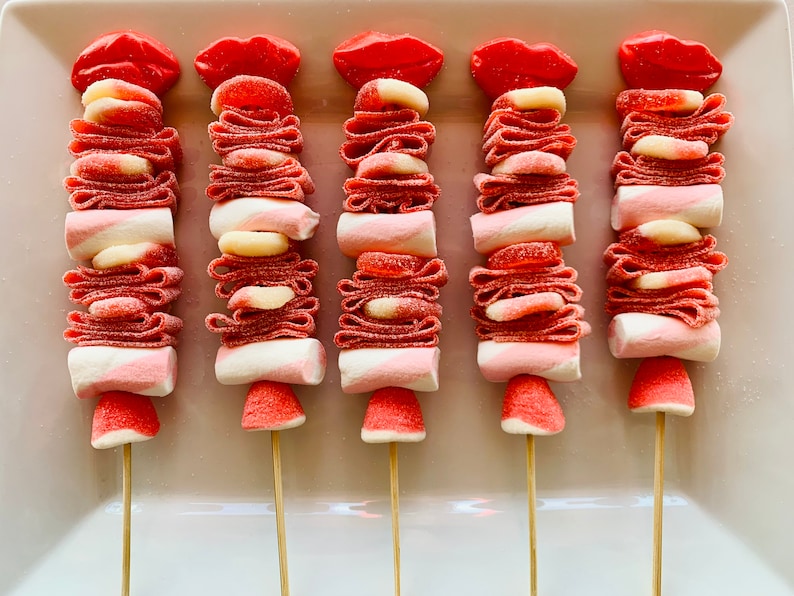Op de afbeelding: Vijf roze en witte snoepspiesjes met rode snoeplippen erop. Elke spies heeft een marshmallow, roze en witte snoepjes en een roze snoepje in de vorm van een hart.