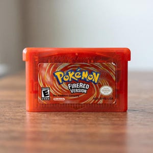 Könnte beinhalten: Ein durchscheinendes rotes Game Boy Advance Videospielmodul für Pokémon FireRed Version. Das Etikett zeigt einen wirbelnden orangefarbenen und roten Hintergrund mit dem blauen und gelben Pokémon-Logo, dem Text FireRed Version, einer ESRB E-Bewertung und dem Nintendo-Logo.
