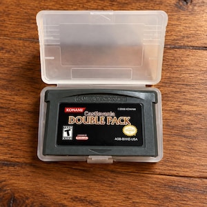 May include: A grey Game Boy Advance video game cartridge titled Castlevania Double Pack, featuring a black label with white and gold text, a Konami logo, and a Teen ESRB rating, housed inside a clear, hinged plastic protective storage case.