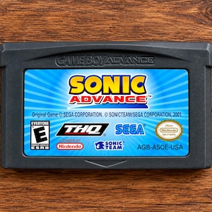 May include: A dark grey Game Boy Advance game cartridge for Sonic Advance. The label features a blue sunburst background with the title Sonic Advance in yellow and red text. It includes logos for THQ, Sega, Sonic Team, Nintendo, and the ESRB E rating.