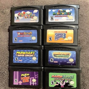 May include: A collection of eight black Game Boy Advance game cartridges featuring titles including Super Mario Advance, Super Mario World, Yoshi's Island, Super Mario Bros 3, Mario Kart Super Circuit, Mario Party Advance, Mario vs Donkey Kong, and Mario and Luigi Superstar Saga.