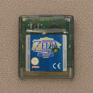 May include: A translucent green Game Boy Color video game cartridge for The Legend of Zelda: Oracle of Ages. The label features blue wood-grain texture, the game title, a Nintendo seal of quality, and the text CGB-AZ8P-EUR, Made in Japan.