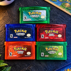 May include: Five Game Boy Advance video game cartridges including Pokemon Emerald, Sapphire, Ruby, FireRed, and LeafGreen versions. Each cartridge features a translucent colored plastic shell and a label with the game title and ESRB rating.