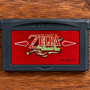 May include: A dark grey Game Boy Advance video game cartridge with a red label featuring the title The Legend of Zelda The Minish Cap, a sword illustration, and the text AGB-BZME-USA printed on the front of the plastic casing.