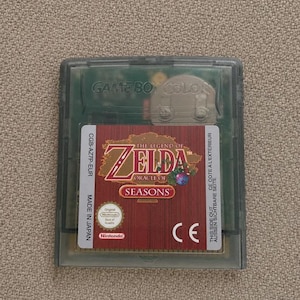 May include: A translucent grey Game Boy Color game cartridge for The Legend of Zelda: Oracle of Seasons. The label features a wood-grain background, the game title, a Nintendo Seal of Quality, and the text CGB-AZ7P-EUR, Made in Japan, and CE.