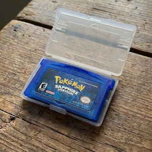 May include: A blue Game Boy Advance video game cartridge titled Pokémon Sapphire Version, featuring an E rating symbol and the text AGB-AXPE USA, housed inside a clear, rectangular, hinged plastic protective storage case.
