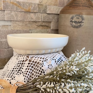 May include: An oval-shaped, off-white ceramic bowl with a smooth interior and a fluted, scalloped exterior base. The bowl has a matte finish and sits on a white crocheted lace doily, positioned in front of a large, tan-coloured stoneware jug.