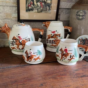 Peut inclure: Un service à thé en céramique comprenant une théière, un pot à lait et deux tasses, tous décorés de scènes en relief représentant des cavaliers à cheval et des chiens de chasse. Les pièces sont crème avec des détails peints en rouge, marron et vert.