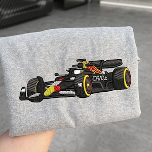 Może przedstawiać: Szary melanżowy t-shirt ze szczegółowym haftem przedstawiającym czarny bolid Formuły 1. Samochód posiada żółte i czerwone akcenty na karoserii oraz oponach, a na boku podwozia wyhaftowano biały napis ORACLE.