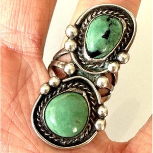May include: A silver ring featuring two teardrop-shaped green stones with dark inclusions. The stones are set in twisted wire bezels and accented with small silver spheres. The band is constructed from multiple silver wires that connect the settings.