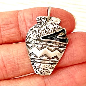 May include: A silver-toned metal pendant shaped like a small vase or amphora. The surface features a textured, speckled finish on the upper and lower sections, with a smooth middle band decorated with two horizontal rows of engraved zigzag patterns.