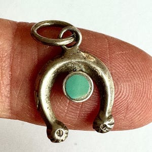 May include: A silver-toned metal horseshoe charm featuring a small, round, flat turquoise-colored inlay centered within the arch. The charm includes a circular jump ring at the top for attachment and has textured, grooved detailing at the ends.
