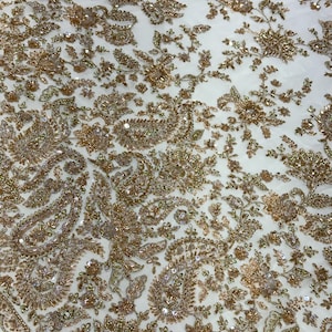 Può includere: Un tessuto bianco con intricati ricami dorati e paillettes disposti in densi motivi paisley e floreali. Il design copre la superficie con fili metallici, piccole perline e paillettes riflettenti, creando un aspetto strutturato.