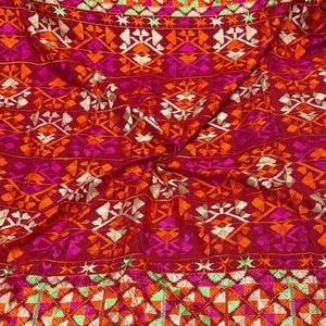 Könnte beinhalten: Ein tiefroter Stoff mit aufwendigen Stickereien in leuchtenden Farben wie Orange, Pink, Weiß und Limettengrün. Das Textil zeigt ein sich wiederholendes geometrisches Muster aus Dreiecken und stilisierten Blumenmotiven auf der gesamten Fläche.