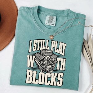 May include: A teal short-sleeved t-shirt featuring a graphic of a car engine and the text I STILL PLAY WITH BLOCKS in cream-colored block lettering. The shirt has a crew neckline and a Comfort Colors brand label printed on the inside collar.