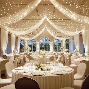 May include: A banquet hall featuring round tables covered in white tablecloths, surrounded by white chairs. The ceiling is draped with sheer white fabric and illuminated by strings of warm white fairy lights, creating a soft, glowing atmosphere.