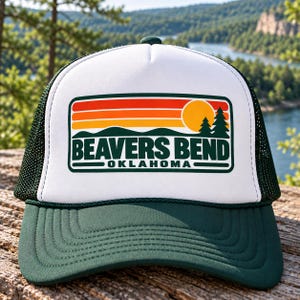 May include: A white and dark green trucker hat featuring a rectangular graphic with a sunset, pine trees, and the text BEAVERS BEND OKLAHOMA. The cap has a solid white front panel, a dark green brim, and a dark green mesh back with an adjustable snap.