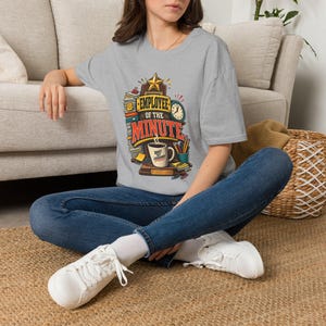 Puede incluir: Una camiseta gris jaspeado con un gráfico de una placa de premio de madera con una estrella dorada, un reloj, pilas de papel, lápices y una taza de café. El texto dice "EMPLOYEE OF THE MINUTE" y "BRIEF BUT LEGENDARY" en la taza de café.