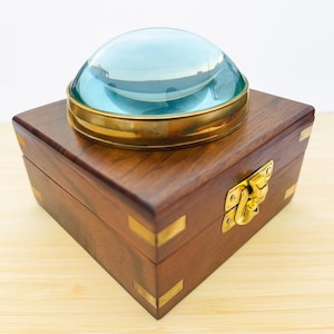Può includere: Una scatola quadrata in legno con inserti in ottone agli angoli e una chiusura in metallo dorato. Sopra la scatola poggia una lente d'ingrandimento circolare con una spessa cornice in ottone e una lente in vetro bombata color azzurro.