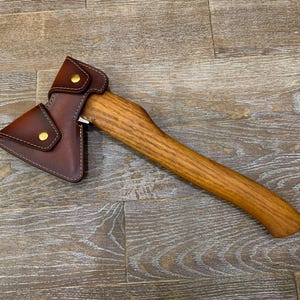 Puede incluir: Un hacha de mano con un mango de madera curvado con veta visible y una funda de cuero marrón que cubre la cabeza de metal. La funda está asegurada con dos broches de metal color latón y presenta costuras visibles a lo largo de los bordes.