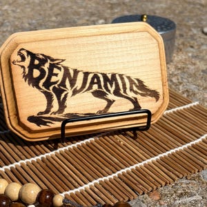 May include: A rectangular light-colored wooden plaque featuring a dark wood-burned illustration of a howling wolf. The name BENJAMIN is integrated into the body of the wolf in bold, dark lettering. The plaque is displayed on a black metal stand.