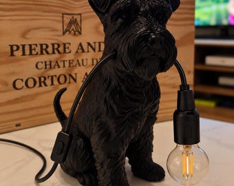 May include: A matte black table lamp shaped like a seated Schnauzer dog. The dog holds a black electrical cord in its mouth, which extends to a black socket holding a clear glass Edison-style light bulb with a visible vertical filament inside.