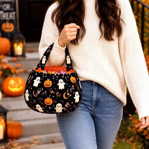 May include: A black fabric trick-or-treat bag featuring a repeating pattern of white ghosts, orange pumpkins, and small crescent moons. The bag has a rounded shape with a matching fabric handle and a bright orange interior lining visible at the top.