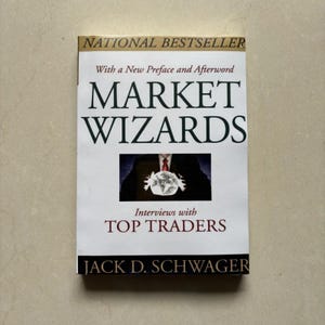 May include: A paperback book titled Market Wizards: Interviews with Top Traders by Jack D. Schwager. The white cover features black text, a gold banner at the top, and a central image of a person in a suit holding a glowing crystal ball.