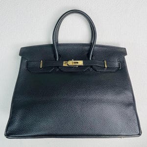 Op de afbeelding: Een zwarte leren handtas met een getextureerde korrelige afwerking, voorzien van twee afgeronde handvatten en een goudkleurige metalen draaisluiting in het midden van de voorflap. De tas heeft een gestructureerde trapeziumvorm.