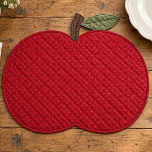 May include: A quilted fabric placemat shaped like a red apple with a brown stem and a green leaf. The red fabric features a subtle tonal pattern and diagonal stitching lines across the surface. The placemat is designed with a rounded apple silhouette.