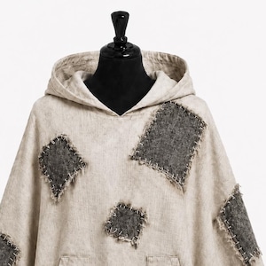 May include: A beige hooded poncho featuring dark gray rectangular patches with visible white cross-stitching along the edges. The garment includes two front pockets and a pointed hemline, constructed from a textured fabric with a natural finish.