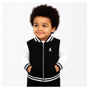 May include: A black and white varsity jacket featuring a full-length silver zipper, white sleeves, and a white striped ribbed collar, cuffs, and hem. The jacket displays a white letter A embroidered on the left chest area.