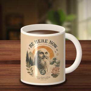 Op de afbeelding: Een witte keramische mok met een beige grafische print van Ram Dass met een baard. De tekst luidt BE HERE NOW, THERE IS NO OTHER MOMENT, en We're all just walking each other home. RAM DASS. Bevat illustraties van bergen, bomen, een lotusbloem en een Om-symbool.