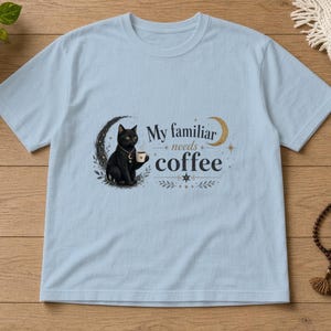 May include: A light blue short-sleeve t-shirt featuring a graphic of a black cat holding a coffee mug, positioned within a crescent moon. The text reads, My familiar needs coffee, accompanied by small star and leaf illustrations.