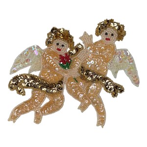 Puede incluir: Un parche decorativo que muestra dos querubines elaborados con cuentas de color melocotón y lentejuelas doradas. Las figuras tienen alas iridiscentes, fajas de lentejuelas doradas y uno sostiene una pequeña flor de cuentas rojas y verdes.