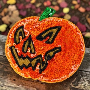 Puede incluir: Un objeto decorativo con forma de calabaza cubierto de lentejuelas de color naranja brillante. Presenta una cara de jack-o'-lantern delineada con cuentas negras y detalles dorados en los ojos, la nariz y la boca dentada. Un tallo de lentejuelas verdes corona la parte superior.