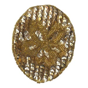 May include: An oval-shaped patch featuring a central floral motif crafted from gold-toned beads and metallic sequins. The surrounding area is densely covered in rows of shimmering silver-toned sequins, creating a textured and reflective surface.