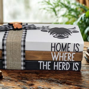 May include: A stack of three decorative wooden blocks painted white, brown, and black. The top block features a black highland cow graphic. The text reads "HOME IS WHERE THE HERD IS" across the blocks. A burlap and black-and-white gingham ribbon wraps around the stack, topped with a small wooden highland cow cutout.