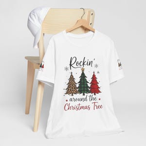 Può includere: Una t-shirt bianca a maniche corte con la grafica di tre alberi di Natale con stampa leopardata, motivo a quadri verde e rosso. Il testo Rockin' around the Christmas Tree è scritto in nero e rosso. È visibile anche un berretto da baseball bianco.