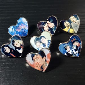 May include: A collection of seven heart-shaped metal pins, each featuring a different photographic image of musicians. The pins display various band members in performance or portrait settings, with silver-toned metal edges framing each heart.