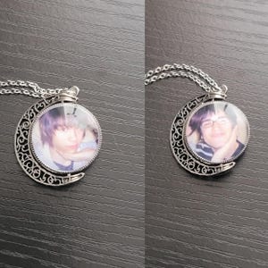 May include: A silver-coloured pendant necklace featuring a crescent moon frame with intricate filigree cutouts. The circular centre displays a photographic portrait of a person. The necklace includes a delicate silver chain attached to the top.