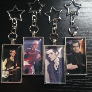 May include: Four rectangular keychains featuring silver-toned star-shaped rings and metal frames. Each keychain displays a different photographic portrait of a musician, including individuals playing a bass guitar, an electric guitar, and posing.