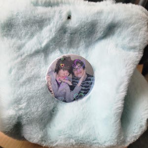 Op de afbeelding: Een lichtblauwe tas van imitatiebont met een ronde speld aan de voorzijde. De speld toont een foto van twee personen met een holografische overlay van kleine gekleurde hartjes en sterretjes verspreid over het oppervlak van de afbeelding.
