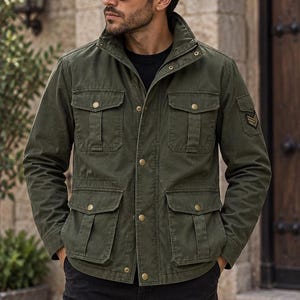 May include: An olive green field jacket featuring a high collar, a front button closure, and four large flap pockets on the front. The jacket includes long sleeves with a chevron patch on the left shoulder and is worn over a plain black shirt.