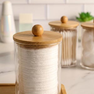 Puede incluir: Un frasco cilíndrico de vidrio transparente lleno con una pila de discos de algodón blancos y redondos. El frasco cuenta con una tapa circular de bambú rematada con un pequeño tirador de madera en forma de bola. Se ven otros dos frascos.