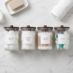 May include: Four clear glass cylindrical canisters with dark brown wooden lids. Each jar features a white square label with black text reading COTTON BALLS, COTTON WOOL PADS, COTTON BUDS, and FLOSS, respectively, containing their labelled items.