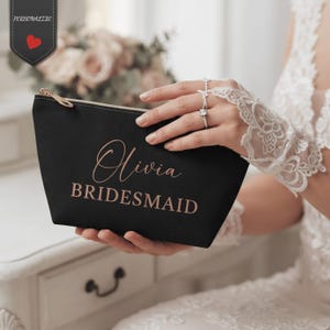 May include: A black zippered pouch featuring the name Olivia and the word BRIDESMAID printed in a metallic rose gold script and block font. The pouch has a gold-toned zipper pull and a small tag in the corner with the text PERSONALIZE.
