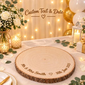 May include: A round wooden slice serving board with natural bark edges, featuring the engraved text "Mr & Mrs Smith" and "16th June 2025" with a small heart symbol. The surface shows visible wood grain rings in a light tan color.